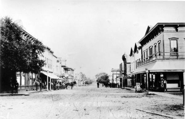 Black and white historical photo of a wide unpaved main street with two story commercial buildings